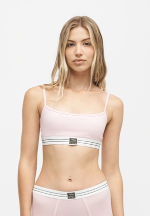 Junge Frau mit langen blonden Haaren trägt ein hellrosa Bralette und passende Unterwäsche mit weißen elastischen Bändern mit der Aufschrift "B Print Underwear."