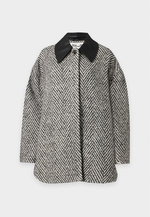 Vrouw draagt een oversized zwart-witte visgraatjas, zwart topje, wijde blauwe jeans en zwarte puntige schoenen, staand tegen een effen achtergrond.