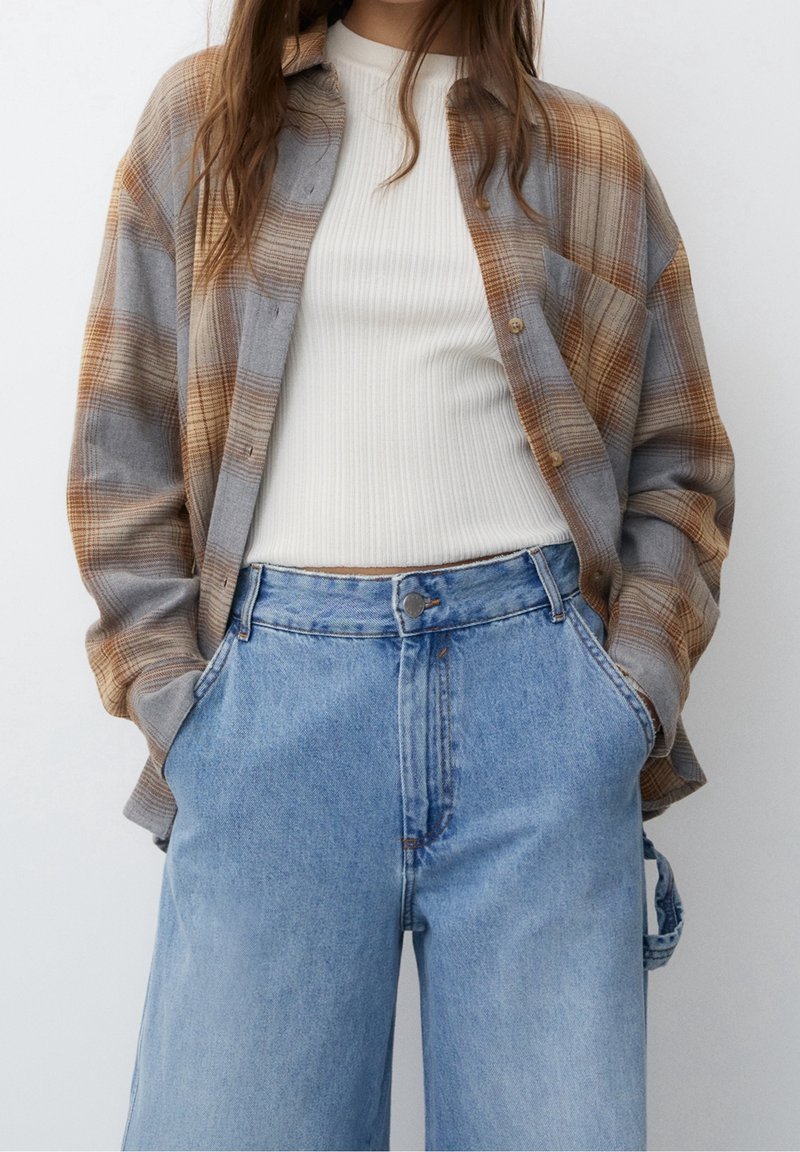 Femme portant un jean taille haute bleu clair avec les mains dans les poches, un haut blanc côtelé et une chemise ouverte à carreaux marron et gris.