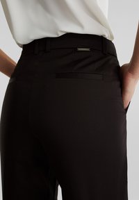 Henkilö, jolla on yllään mustat räätälöidyt housut ja valkoinen sisätasku, yhdellä kädellä taskussa, näyttäen selkävyön yksityiskohtia.