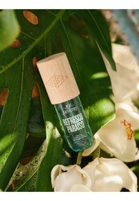 Botella de perfume en spray Refresco Paraíso con tapa de madera, descansando sobre grandes hojas verdes y flores blancas con gotas de agua.