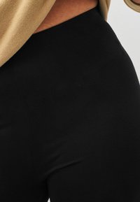 Leggings noirs en tissu lisse et extensible avec une taille haute. La texture visible est mate, sans motifs ni embellissements.