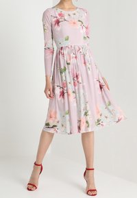 Pitkähihaisessa kukkamekossa on vaaleanpunainen kangas, kerätty vyötärö ja polvenpituinen hame. Lisätty punaisilla korkokengillä.