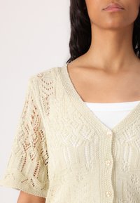 Light yellow knitted cardigan with lace pattern, short sleeves, V-neck, and pearl buttons. Worn over a white tank top.