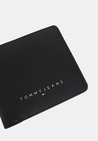 Cartera de cuero negra con el logo en relieve "TOMMYJEANS", forma rectangular, bordes cosidos y sutiles acentos en rojo, blanco y azul.