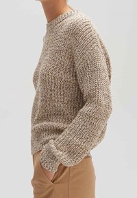 Beżowy i brązowy sweter z melanzowanej dzianiny, z prążkowanymi mankietami i dołem, noszony z beżowymi spodniami, ręka w kieszeni, tors i ramie widoczne z boku.