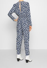 Marinblå blommönstrad jumpsuit med långa ärmar, rynkad midja och nyckelhål i ryggen; kombinerad med vita ankelstövlar och korta, raka ben.