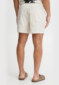 Shorts en coton beige clair avec une taille élastique, comprenant une poche arrière. Le tissu semble lisse et léger.