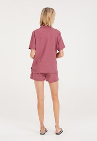 Conjunto de camisa de manga corta con cuello y pantalones cortos en tejido texturizado de color rosa claro, con acentos tonales y un ajuste relajado. Chanclas visibles.
