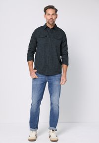Ciemna koszula w wzory z dwoma kieszeniami na piersi, średnio niebieskie dżinsy i jasne trampki. Koszula ma podwinięte rękawy i luźny krój.
