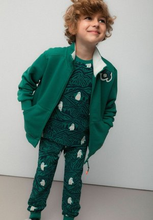 Zielona kurtka na zamek z białym kołnierzem, w połączeniu z wzorzystym topem i spodniami z abstrakcyjnymi wzorami oraz motywami niedźwiedzia polarnego. Miękki materiał.