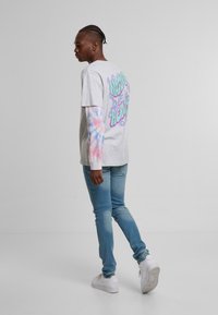 Grå kortermet t-skjorte med fargerik grafikk på ryggen, lagd over en langermet t-skjorte med tie-dye. Lyseblå jeans og hvite sneakers fullfører antrekket.