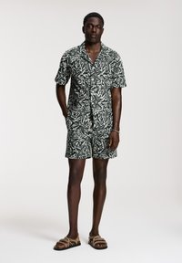 Kurzärmliges, grün-weiß gemustertes Hemd und passende Shorts aus leichtem Stoff, kombiniert mit braunen Sandalen mit Riemen.