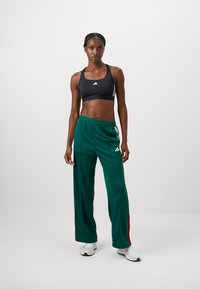 Zwarte sportbeha met wit logo, gecombineerd met groene trainingsbroek met rode zijstrepen en witte accenten. De textuur lijkt soepel en aansluitend.