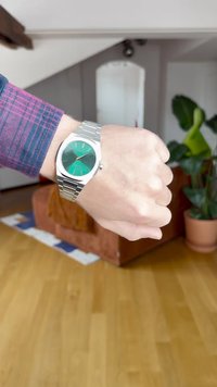 Randmel, millel on hõbedane metallkell, mis näitab 10:09, inimene kannab lilla ruudulist särk, taustal hägune elutuba pruunika diivani ja pott-taimega.