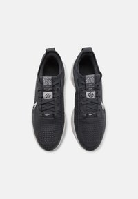 Chaussures de sport noires avec une tige en mesh texturé, fermeture à lacets et accents réfléchissants, dotées d'une semelle grise amortie et d'étiquettes de marque.