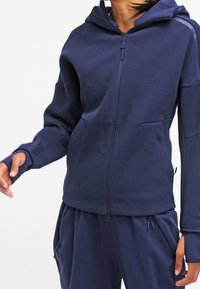 Marinblå zip-up hoodie med fodrad huva, framficka och ribbade muddar. Tillverkad av mjukt material med en slät textur och subtila designdetaljer.