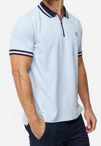 Jasnoniebieska koszulka polo z zamkiem w kołnierzyku, granatowymi akcentami na kołnierzu i rękawach oraz małym haftowanym logo na piersi. Miękka tkanina.