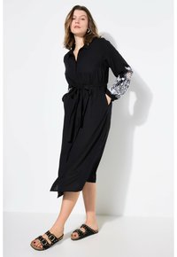 Schwarzes Hemdkleid mit kurzen Ärmeln, das mit weißer floralem Stickerei verziert ist, tailliert mit einem Gürtel und ergänzt durch schwarze Rutschsandalen.