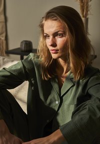 Grønt, struktureret pyjamas sæt består af en knap-op skjorte og bukser, med en slank pasform og subtile mønstre, sat mod en varmtonet baggrund.