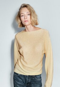 Donna con i capelli corti biondi che indossa un maglione beige e jeans scuri, in piedi su uno sfondo grigio chiaro uniforme.