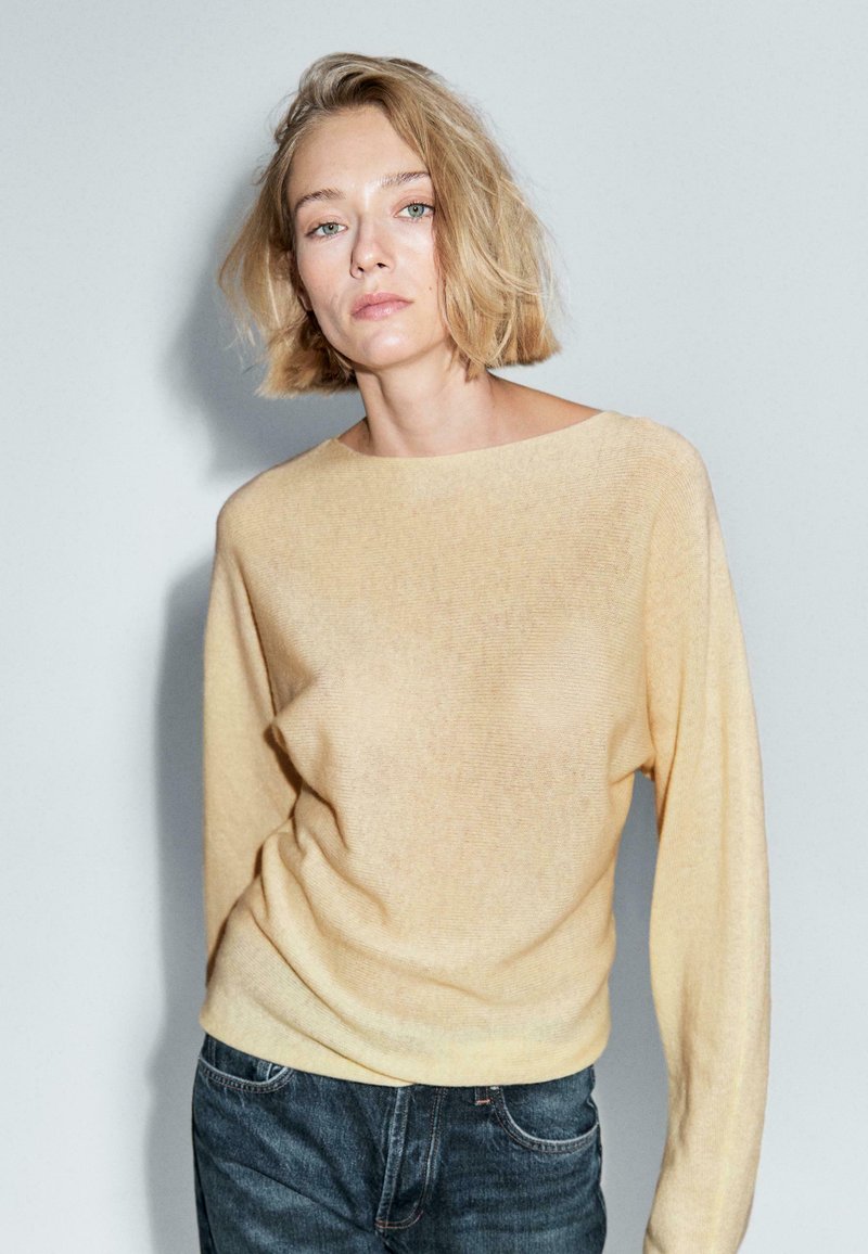 Frau mit kurzem blonden Haar, die einen beigen Pullover und dunkle Jeans trägt, steht vor einem einfarbigen hellgrauen Hintergrund.