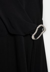 Veselă neagră cu stofă fluidă, având o cataramă texturată și strălucitoare din argint pe mânecă. Suprafață netedă și cădere elegantă.