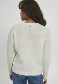 Pull léger beige avec un tricot texturé, col rond et ourlet en côtes. Présente un petit logo au dos. Porté avec un jean bleu.
