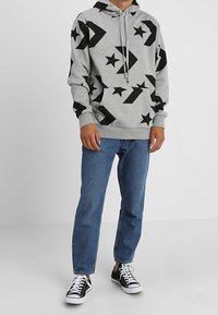 Grey hooded sweatshirt with black star and arrow pattern, drawstring at neckline, paired with blue jeans and black sneakers.