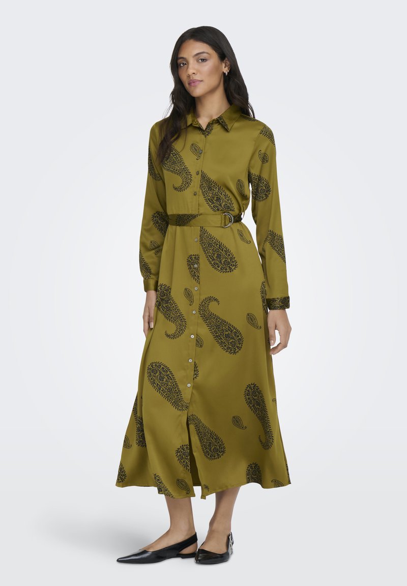 Mujer con un vestido midi de manga larga color verde oliva, abotonado, con patrones de cachemira negros y zapatos planos negros puntiagudos, de pie contra un fondo claro.