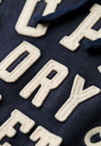 Granatowy materiał z beżowym, szytym napisem. Litery są teksturowane i puszyste, kontrastujące z gładkim tłem. Brak dodatkowych szczegółów.