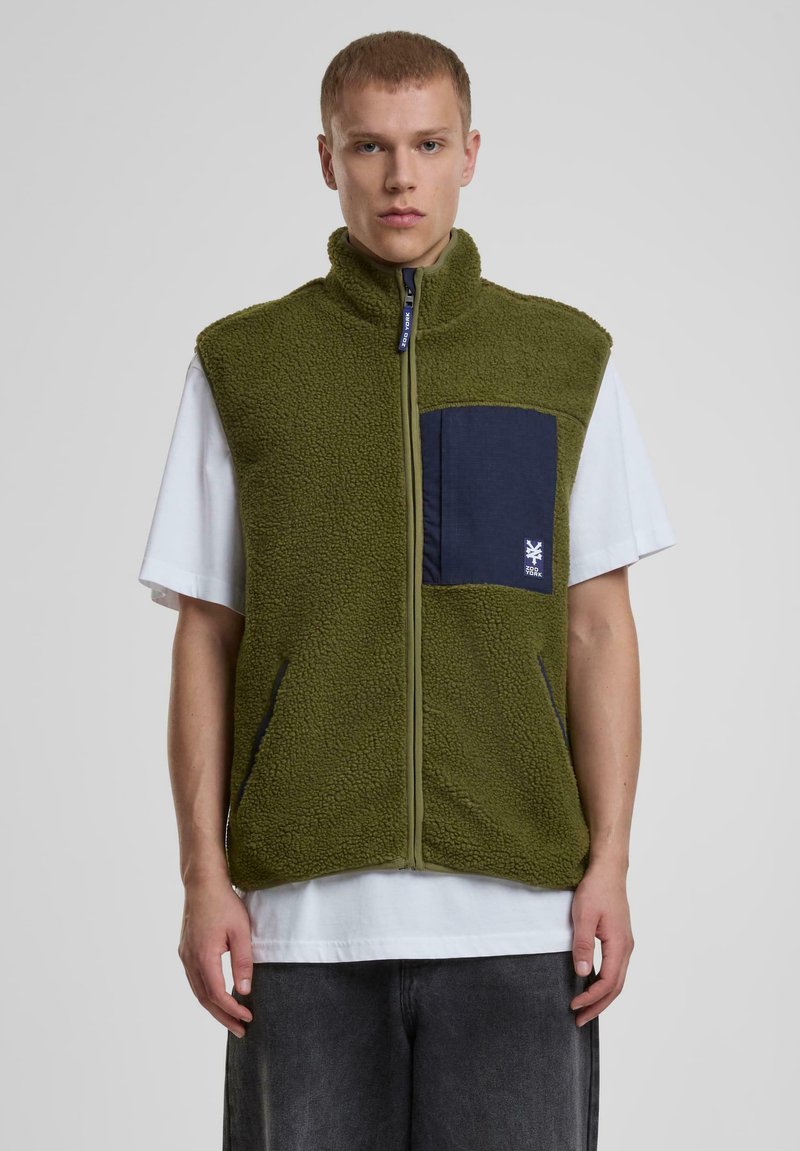 Grüne Fleece-Weste mit hohem Kragen, frontalem Reißverschluss und navyblauer Tasche. Verfügt über strukturierten Stoff und Seitentaschen, über einem weißen Hemd getragen.