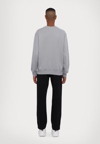 Person som står vänd bort med en grå sweatshirt, svarta byxor och vita sneakers mot en vit enfärgad bakgrund.