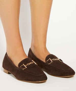 Bruine suède loafers met gouden kettingdetail, gedragen op blote voeten, die de onderbenen en voeten tonen tegen een witte achtergrond.