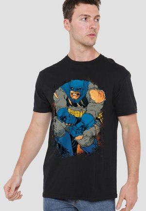 Young man wearing black t-shirt with vintage-style cartoon Batman gripping his knee, looking to the side against plain light background.