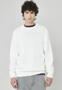 Pull en tricot blanc avec un col rond, présentant une finition texturée et des poignets et un ourlet côtelés, porté par-dessus une chemise sombre.
