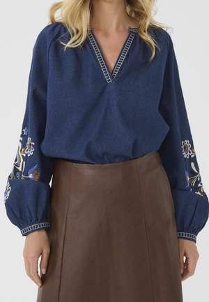 Vrouw die een blauwe blouse met lange mouwen en bloemenborduursel op de mouwen draagt, met een bruine leren rok met hoge taille, poserend tegen een effen achtergrond.