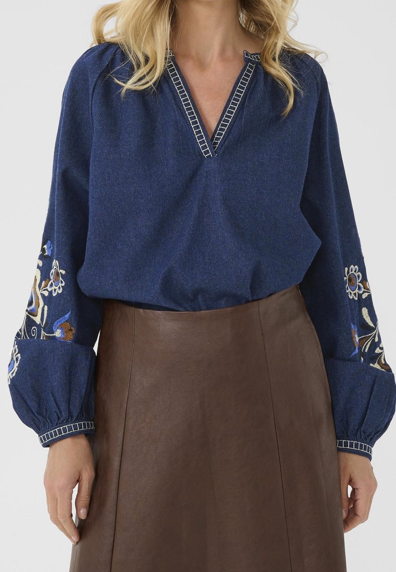 Femme portant un chemisier bleu à manches longues avec broderies florales sur les manches et une jupe en cuir marron taille haute, posant devant un fond uni.