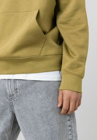 Sweatshirt vert olive avec une poche kangourou et des poignets côtelés, porté sur une chemise blanche. Associé à un jean gris texturé.