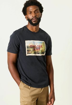Mann mit Bart, der ein schwarzes T-Shirt mit einer Wanderszene und dem Text "Explore More" trägt, Hände in den Taschen, steht vor einem einfarbigen hellen Hintergrund.