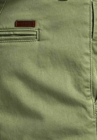 Gros plan sur un pantalon en tissu vert montrant une poche cousue avec une petite étiquette marron de la marque Jack & Jones.