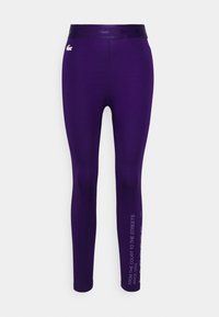 Leggings morados con una banda elástica texturizada. Presentan un logo y un texto blanco que dice "DE LA CANCHA A LAS CALLES" en el lado.