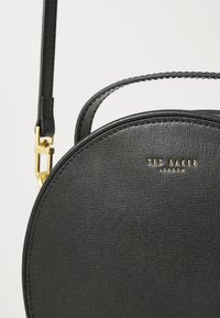 Svart rund handväska med texturerad yta, guldhardware och ett kort handtag. Har en logotyp präglad på framsidan för detaljer.