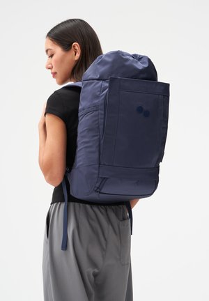 Frau mit einem großen marineblauen Rucksack mit Seitentaschen und einem Reißverschlussfach am Boden, gekleidet in schwarzes Oberteil und graue Hose.