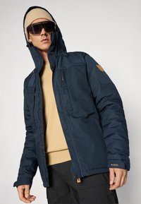 Navy blue insulated jacket with a zip and snap closure, two front pockets, and a hood. Paired with a beige sweater and black pants.