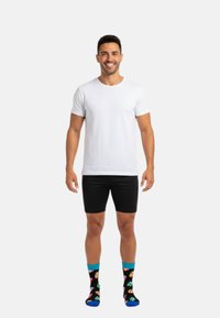 Hvid T-shirt med korte ærmer, sorte shorts og farverige mønstrede sokker med blå kant. Modellen står oprejst mod en ensfarvet baggrund.