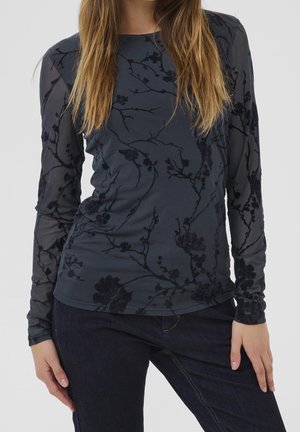 Woman wearing a dark sheer long-sleeve top with black floral patterns and dark blue jeans, standing with hands near hips.