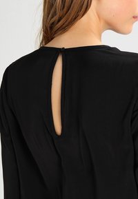 Musta vaate, jossa on takana avaimenreikäleikkaus, pehmeä rakenne ja pitkät hihat, korostaen yksinkertaisia ja tyylikkäitä muotoilutietoja.