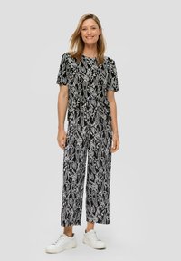 Frau steht in einem passenden Oberteil mit Kurzärmeln und einem schwarz-weißen Muster sowie weit geschnittenen Hosen und weißen Sneakers vor einem einfarbigen Hintergrund.