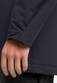 Giacca impermeabile navy con una texture liscia; presenta polsini aderenti e dettagli di cucitura lungo la manica. La mano è parzialmente visibile.